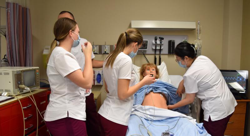 Four nursing students in FON uniforms (white tops and burgandy pants) care for a mannequin in the nursing simulation room
